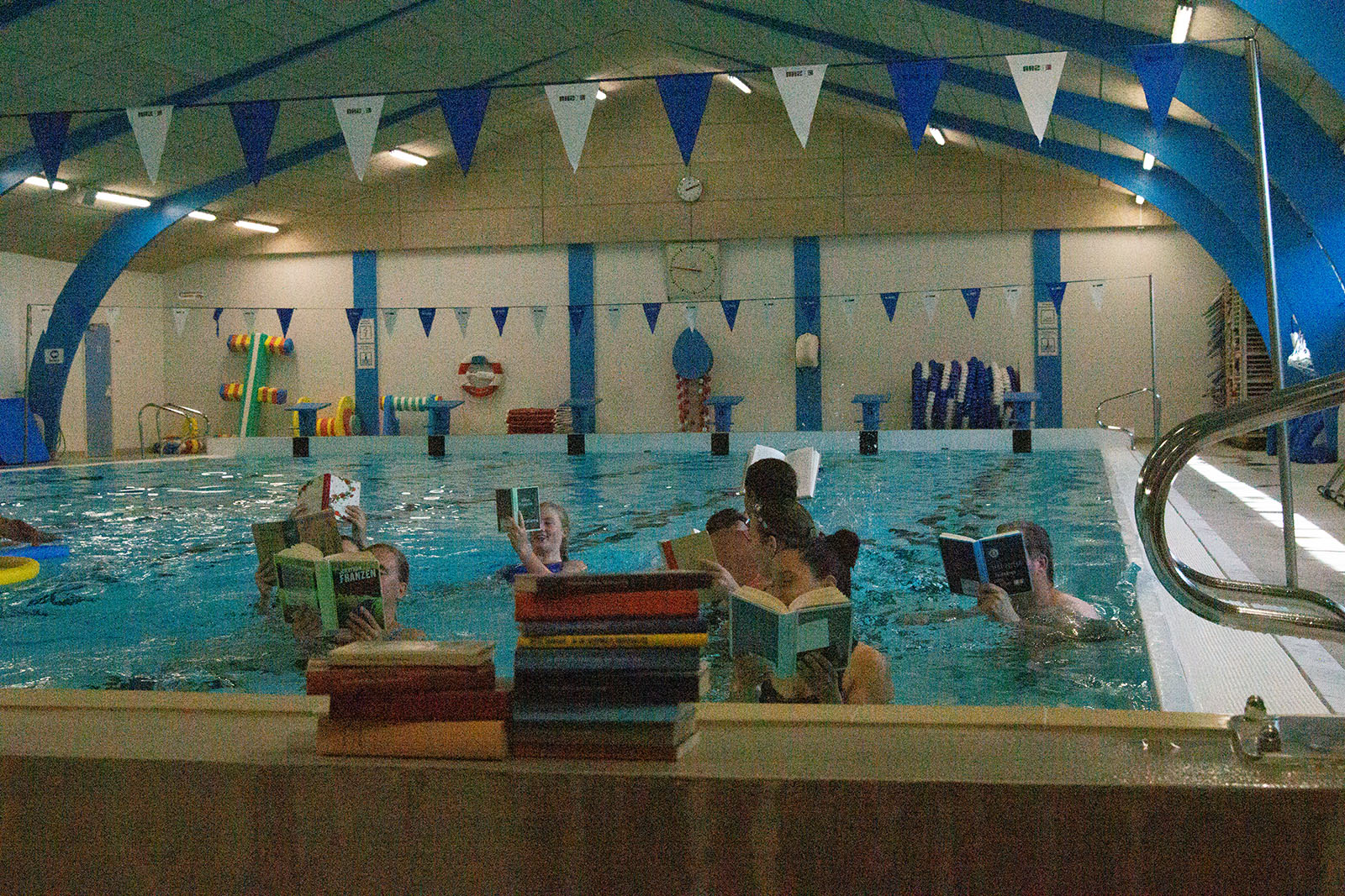 Svøm en bog i Thyregod. En stak af biblioteksbøger på bassinkanten og folk der svømmer og læser i bassinet.